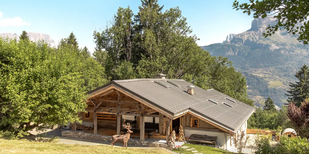 Chalet de Haute Savoie en bois avec terrasse et toit gris, installé dans un jardin arboré, avec des montagnes en arrière-plan sous un ciel bleu.